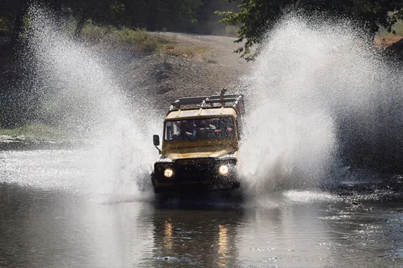 A brown Jeep Safari driving through a river and spraying water.