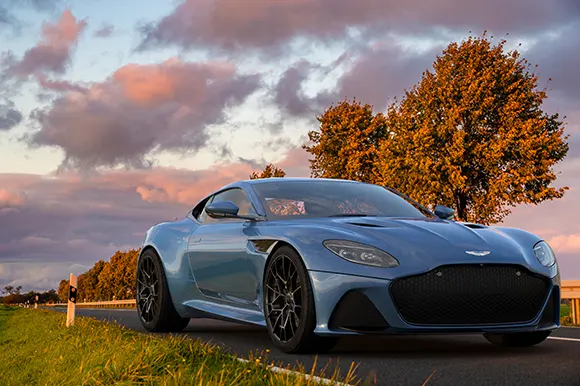 A light blue Aston Martin DBS on a scenic road during golden hour